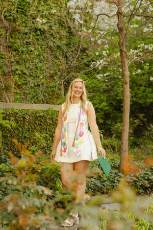 Smiling blonde woman in white dress with colorful sash holding green graduation cap outdoors.