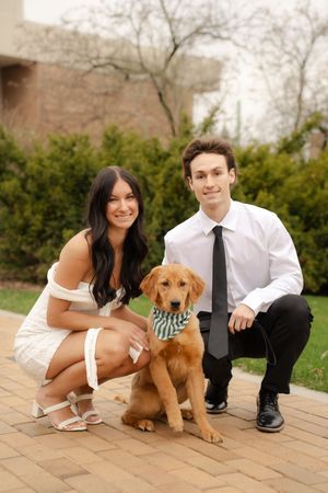 A smiling couple in formal attire crouches beside their golden retriever puppy wearing a bandana outdoors.