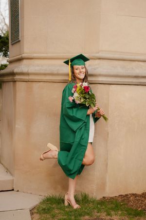 Smiling graduate in green cap and gown holding colorful flower bouquet, posing playfully outside stone building.
