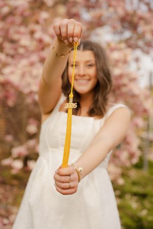 Smiling graduate in white dress holding gold 2026 tassel outdoors with pink cherry blossoms in background.