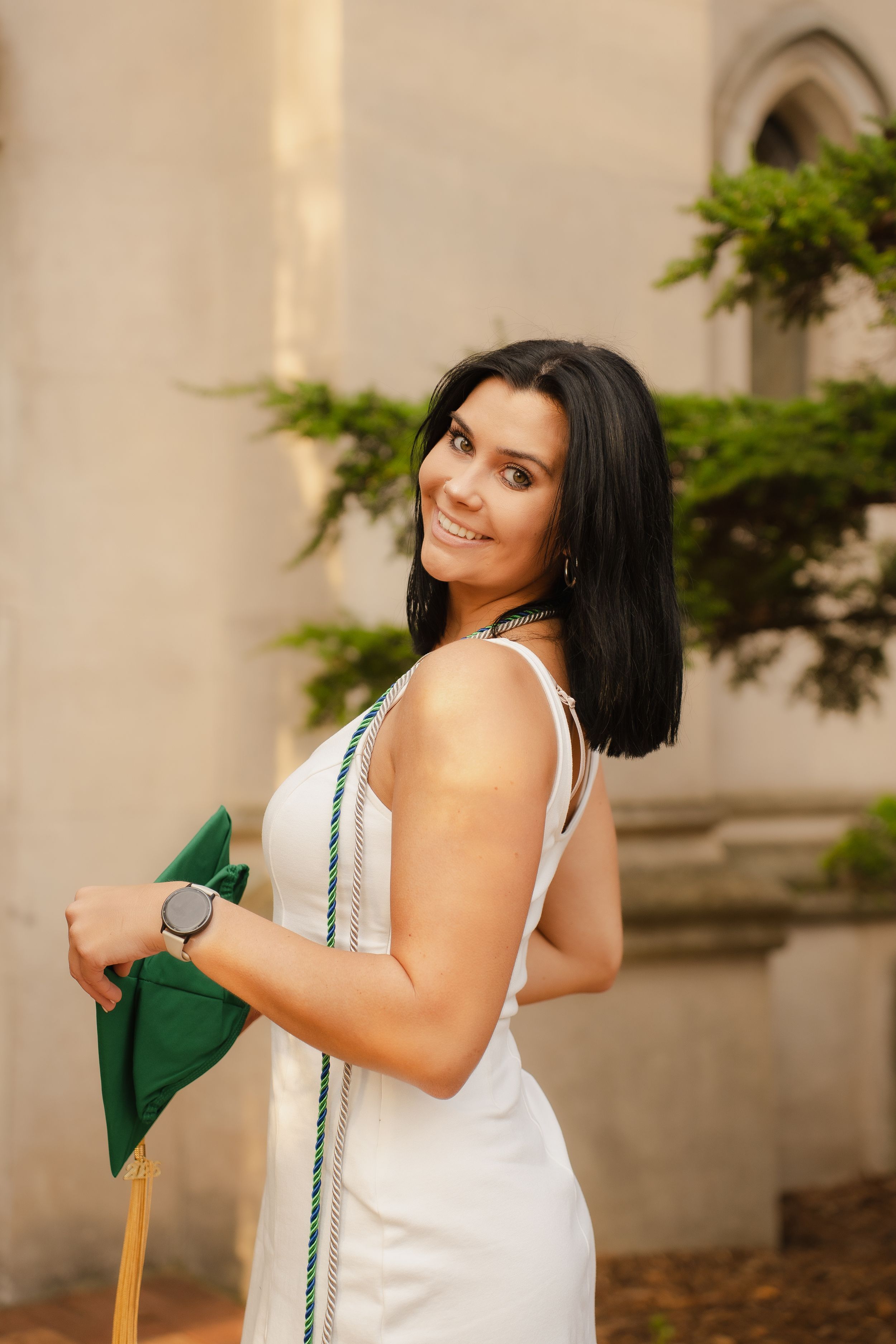 Smiling brunette woman in white dress holding green graduation cap outside historic building.