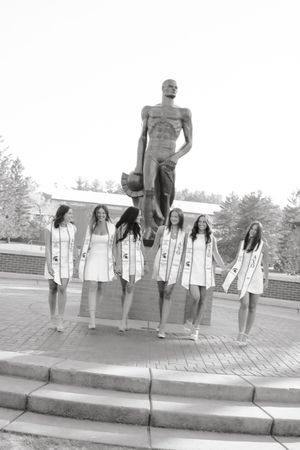 Six female graduates in white dresses and sashes pose before the Spartan Statue at Michigan State University.