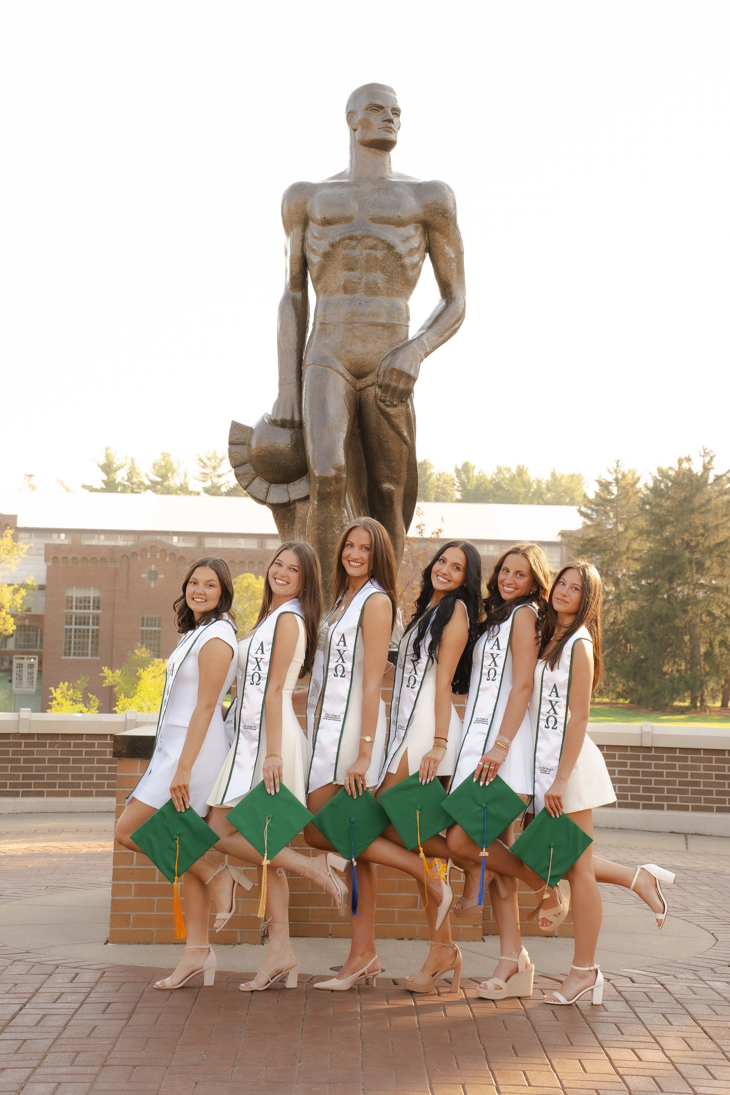 Six graduating women in white dresses and green caps pose in front of the Spartan Statue at Michigan State University.