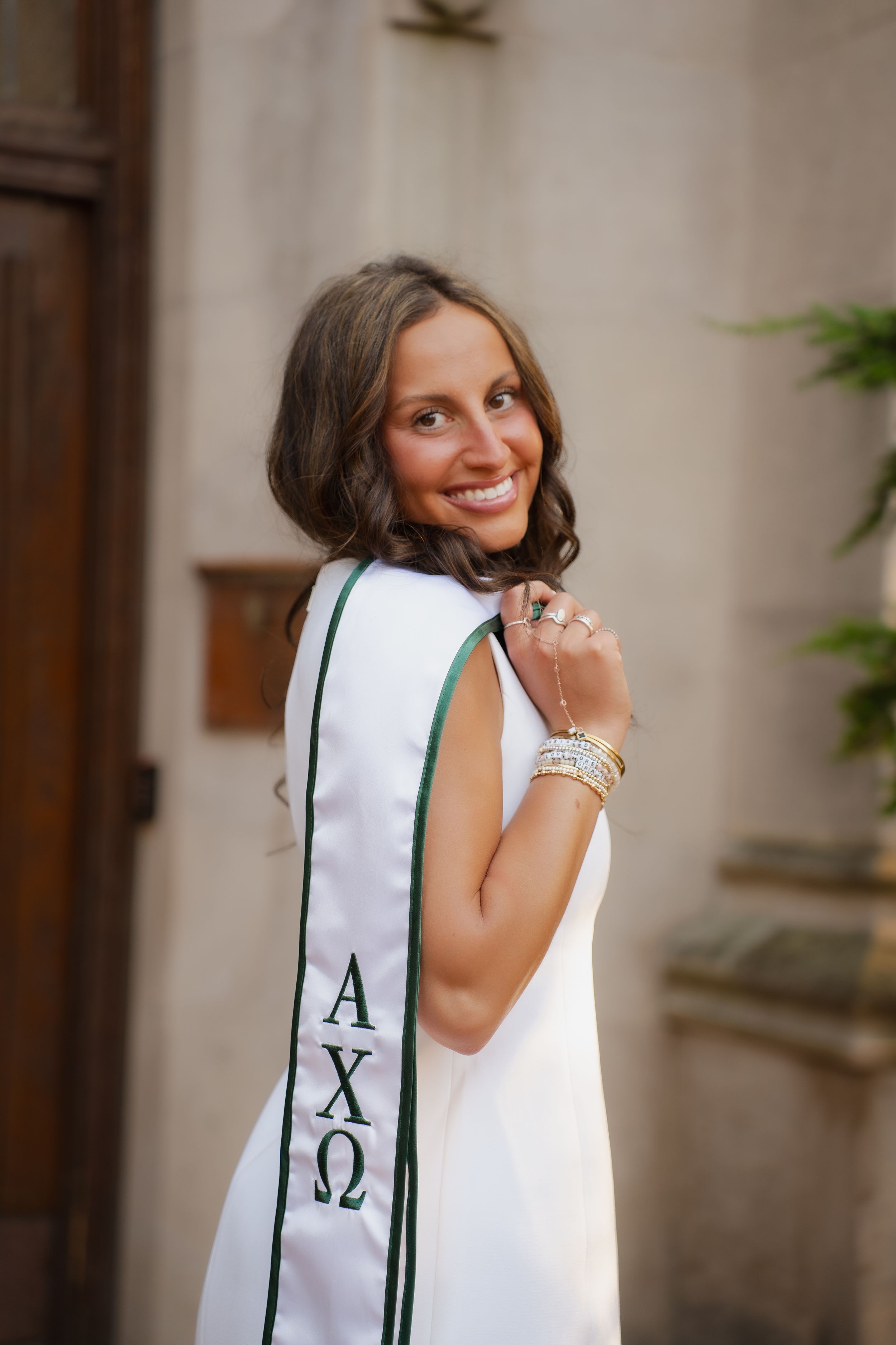 Smiling young woman wearing Alpha Chi Omega graduation stole in white and green, posing outdoors.