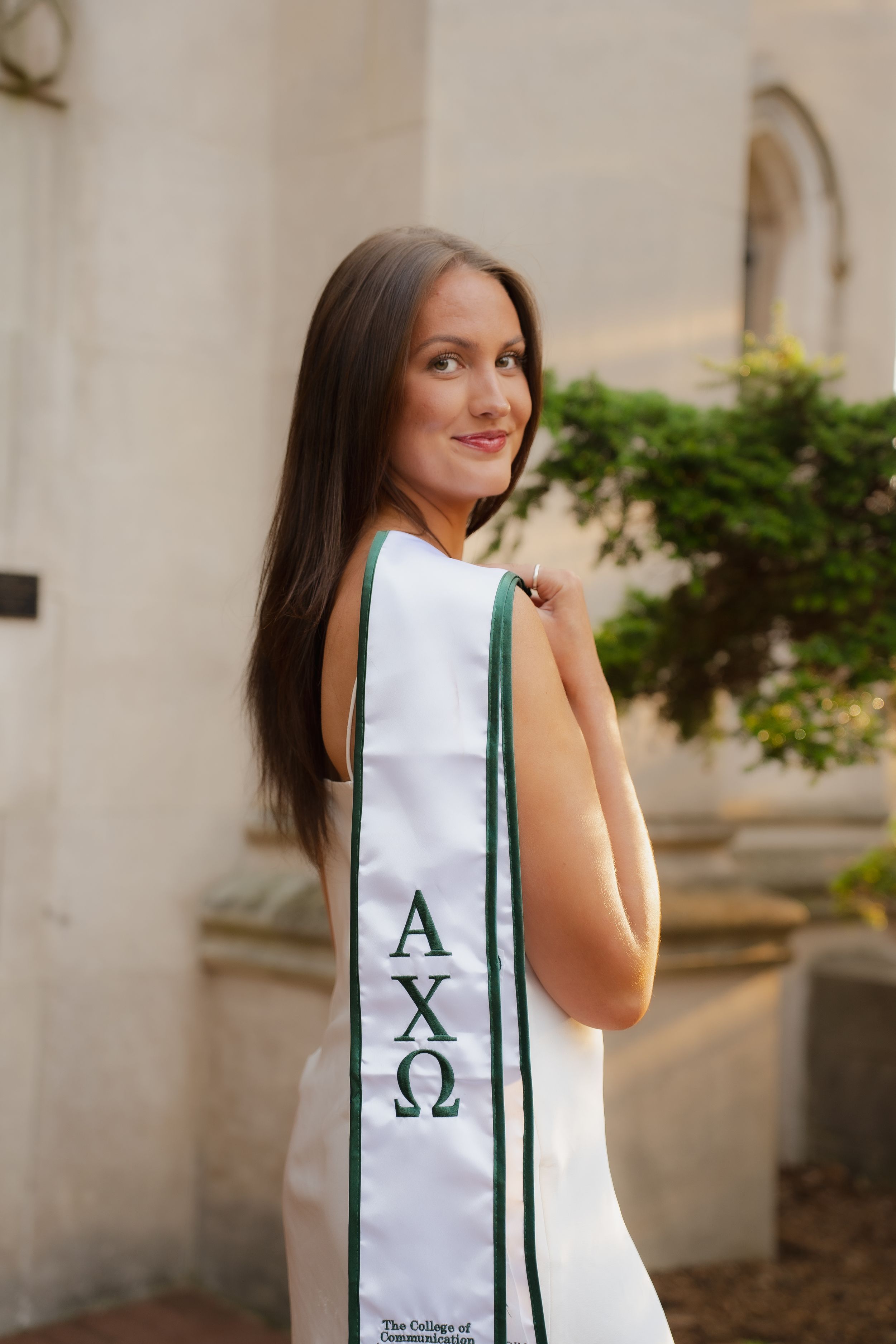 Young woman wearing Alpha Chi Omega Greek sorority graduation sash outdoors near stone building.