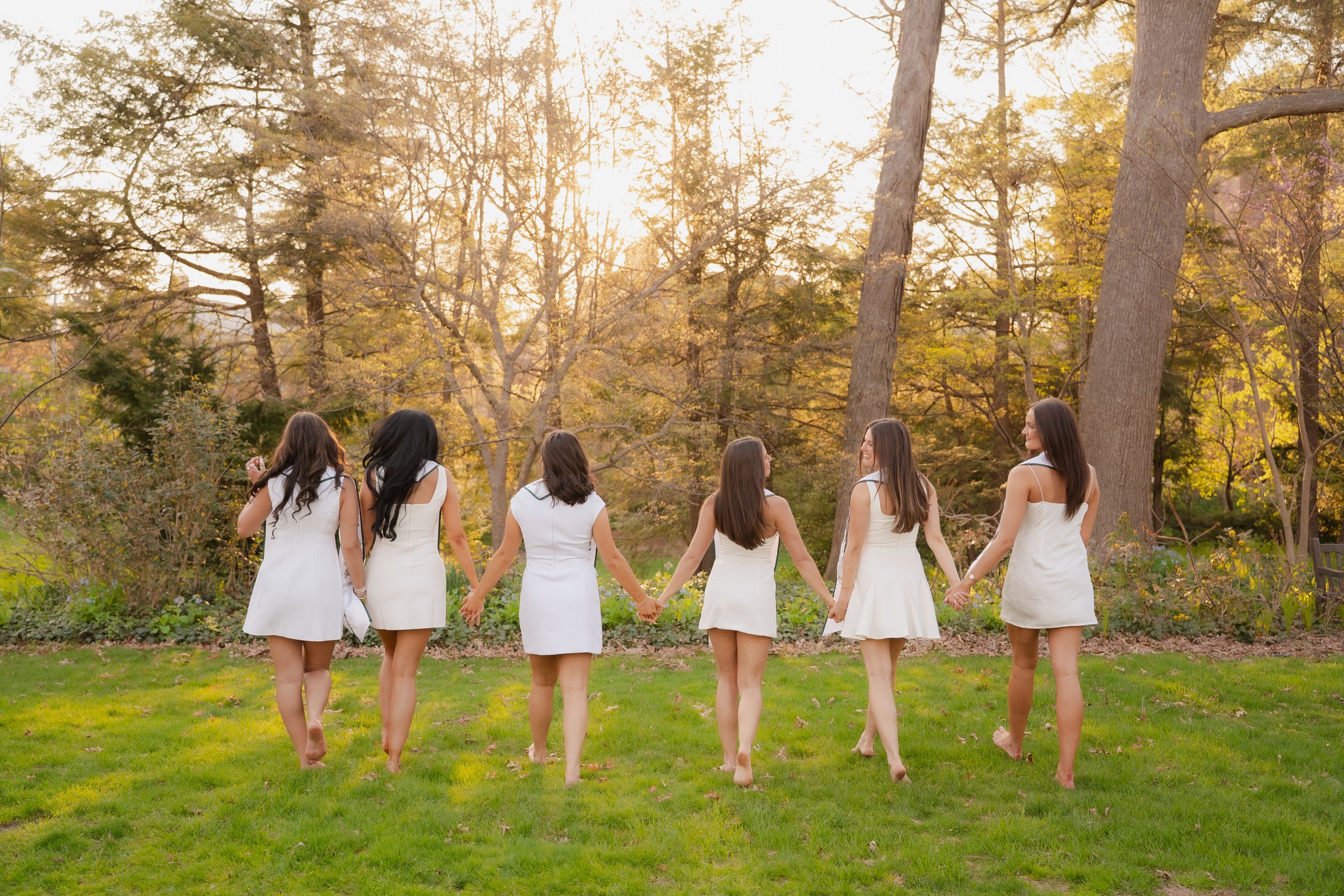 Six women in white dresses hold hands walking through a sunlit garden with lush green grass and autumn trees.