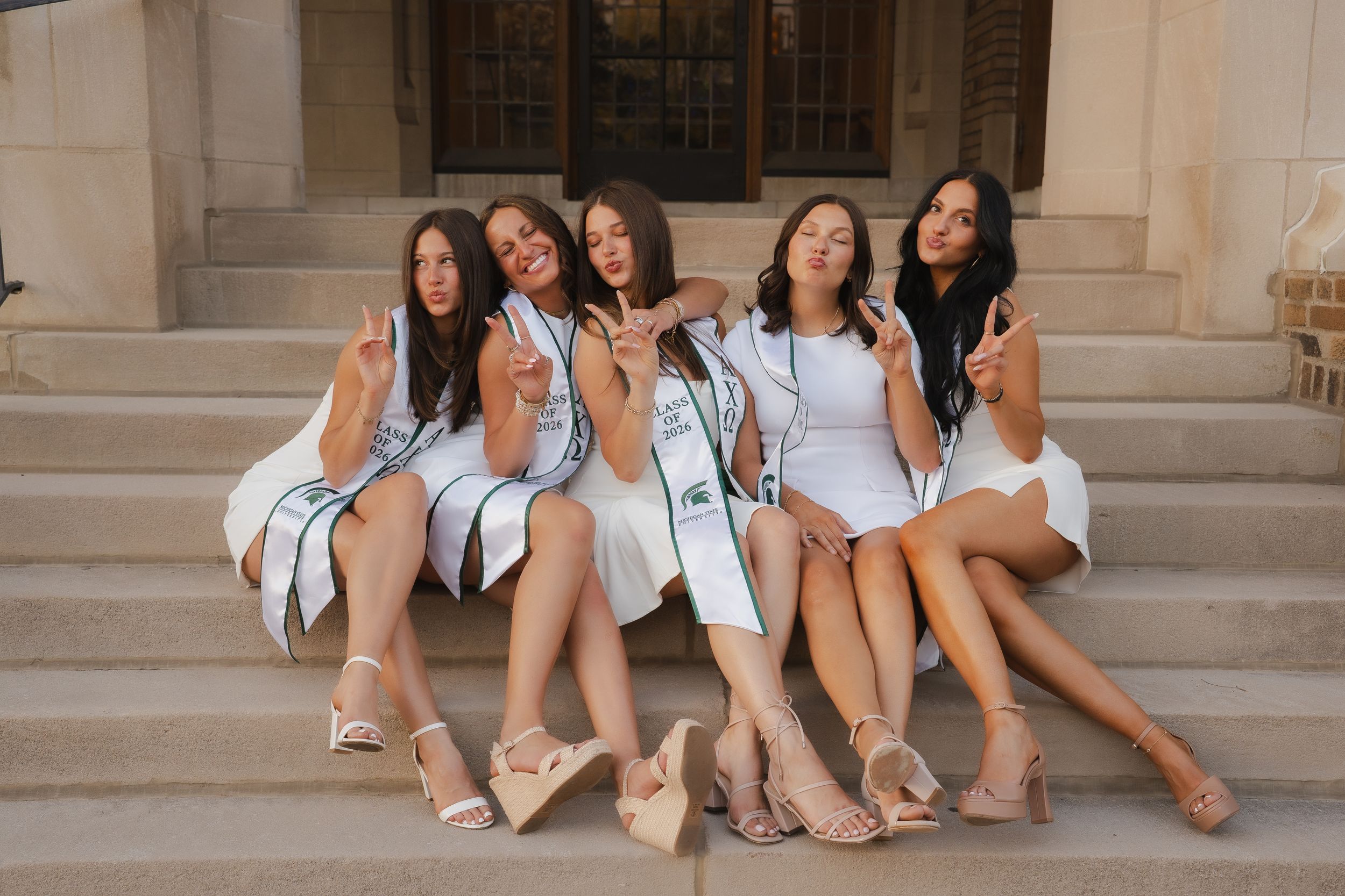Five young women in white dresses and sashes pose on stone steps, making peace signs and duck faces for a photo.