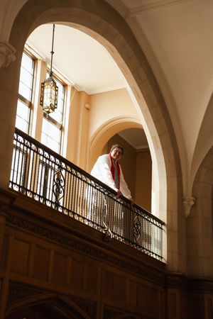 Person in white coat with red scarf leaning on ornate iron balcony railing inside elegant arched building.