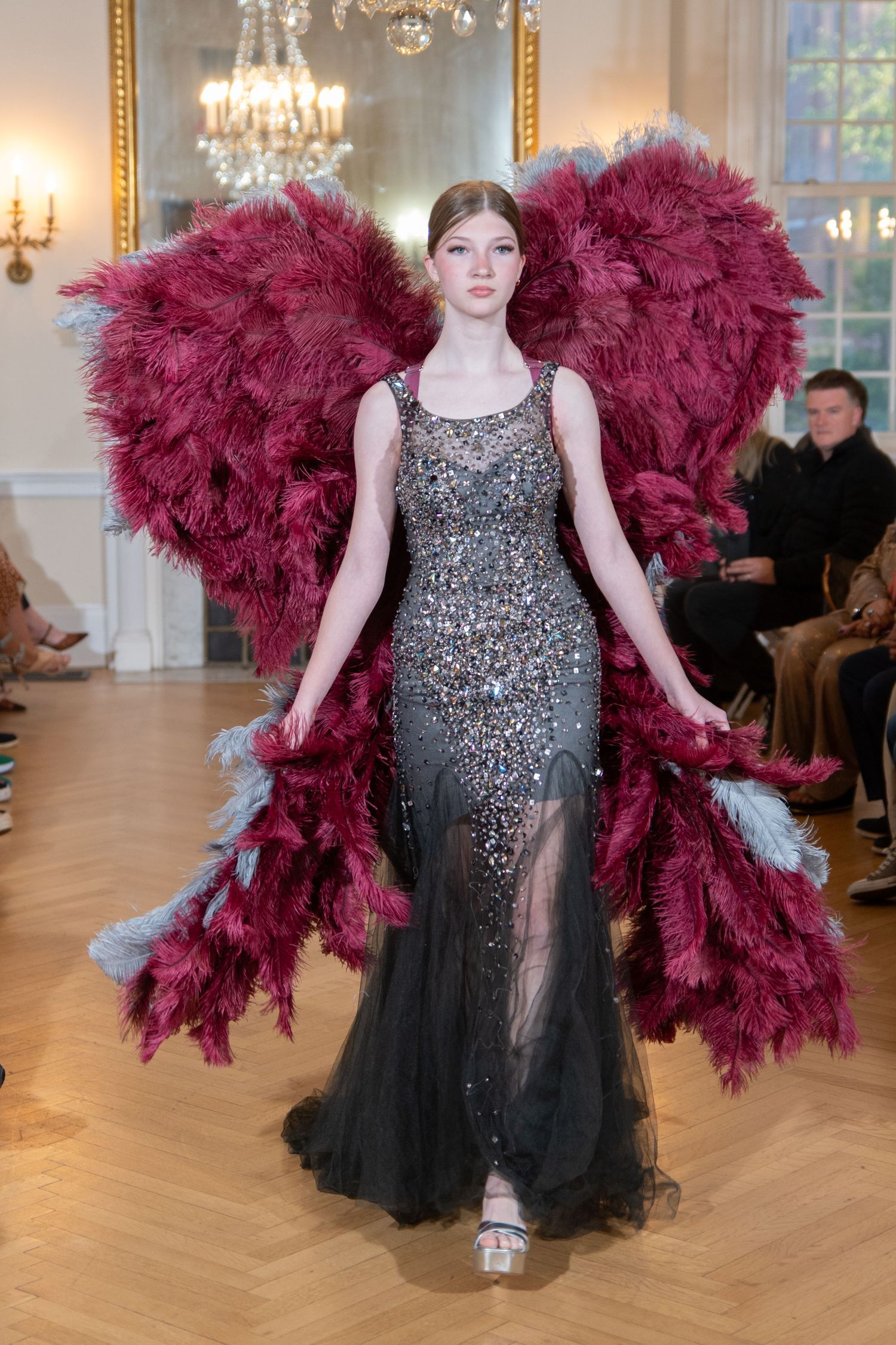 Model wearing beaded gray gown with dramatic crimson feather wings on runway at haute couture fashion show.