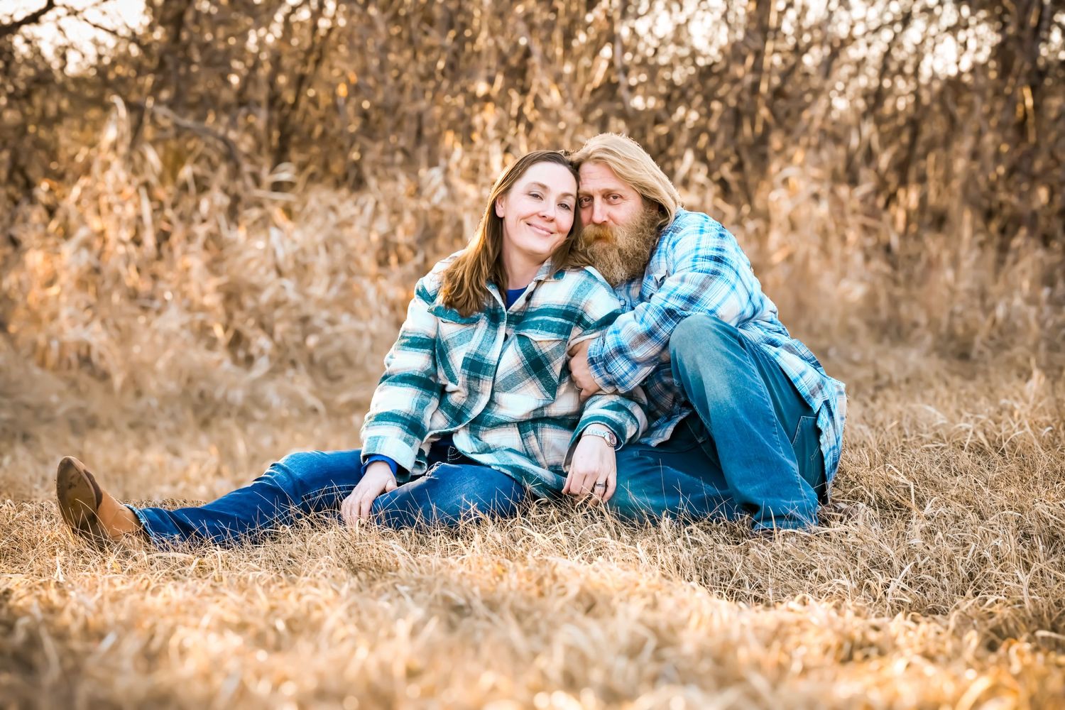 Couple sitting in golden grass field, wearing matching plaid shirts, smiling and embracing in an outdoor autumn setting.