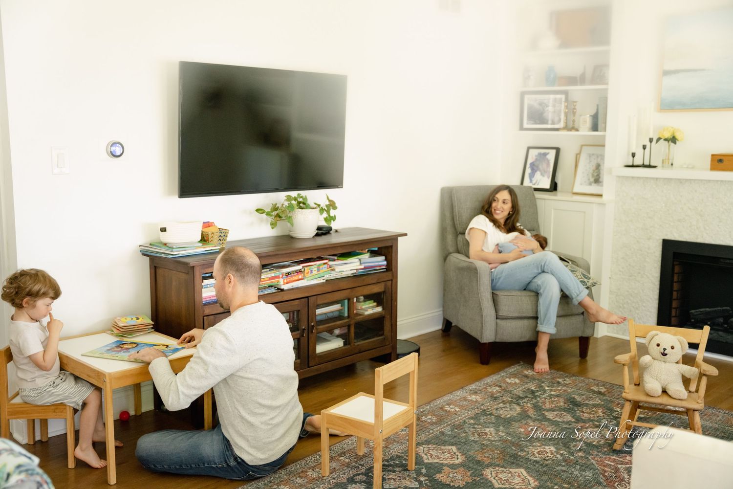 Family relaxing in cozy living room with TV, fireplace, child at table, dad on floor, and mom in armchair.