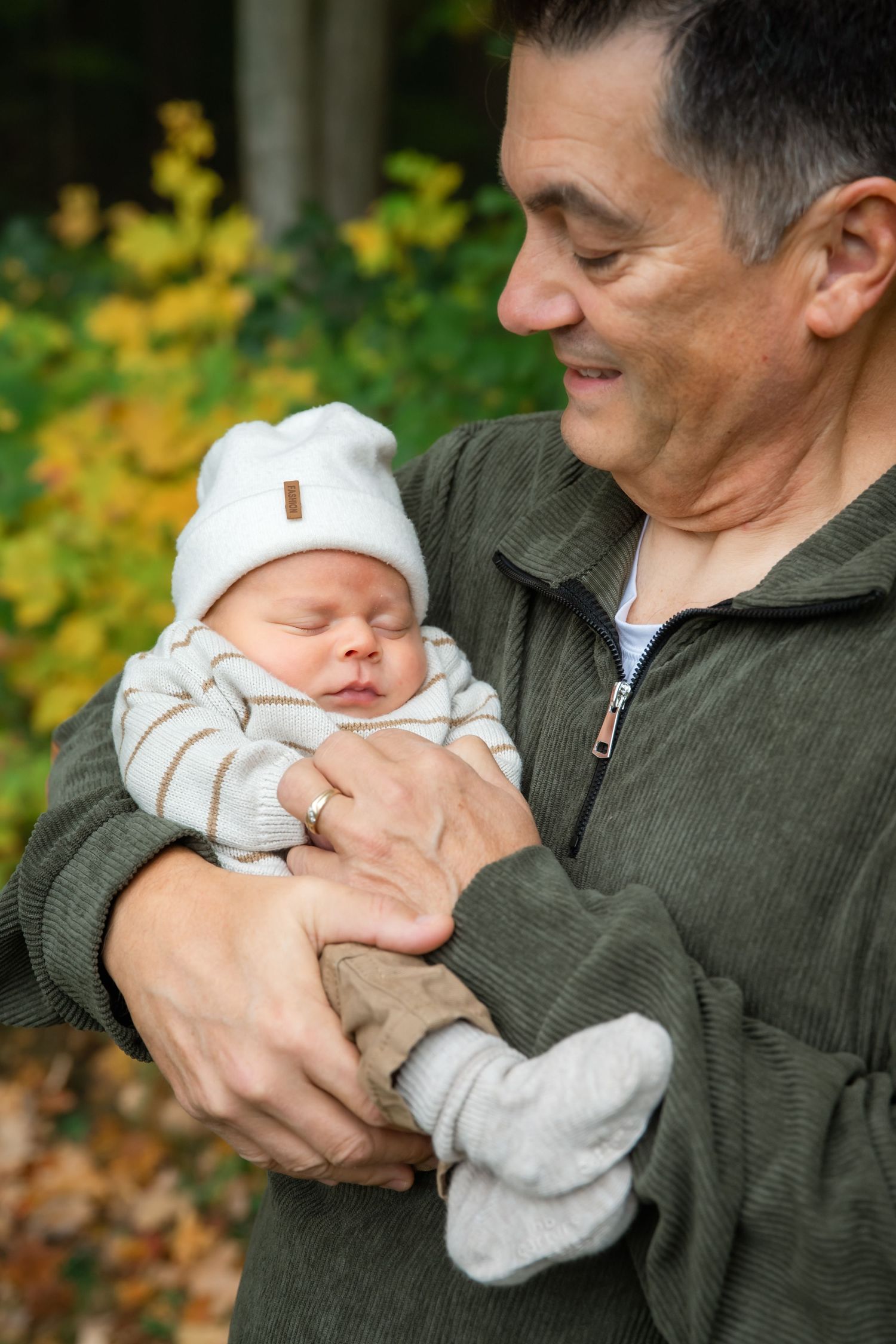 Older man tenderly holding a sleeping newborn baby in a white hat outdoors with autumn foliage background.
