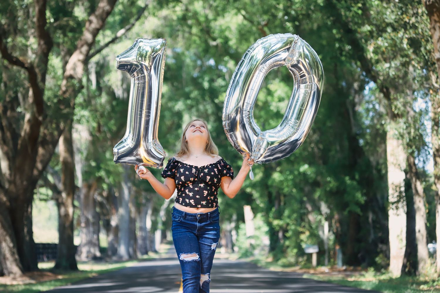 Happy girl celebrates 10th birthday holding large silver number 10 balloons on tree-lined road.