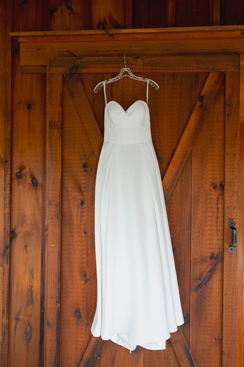 White strapless sweetheart wedding gown hanging on a hanger against rustic wooden barn doors.