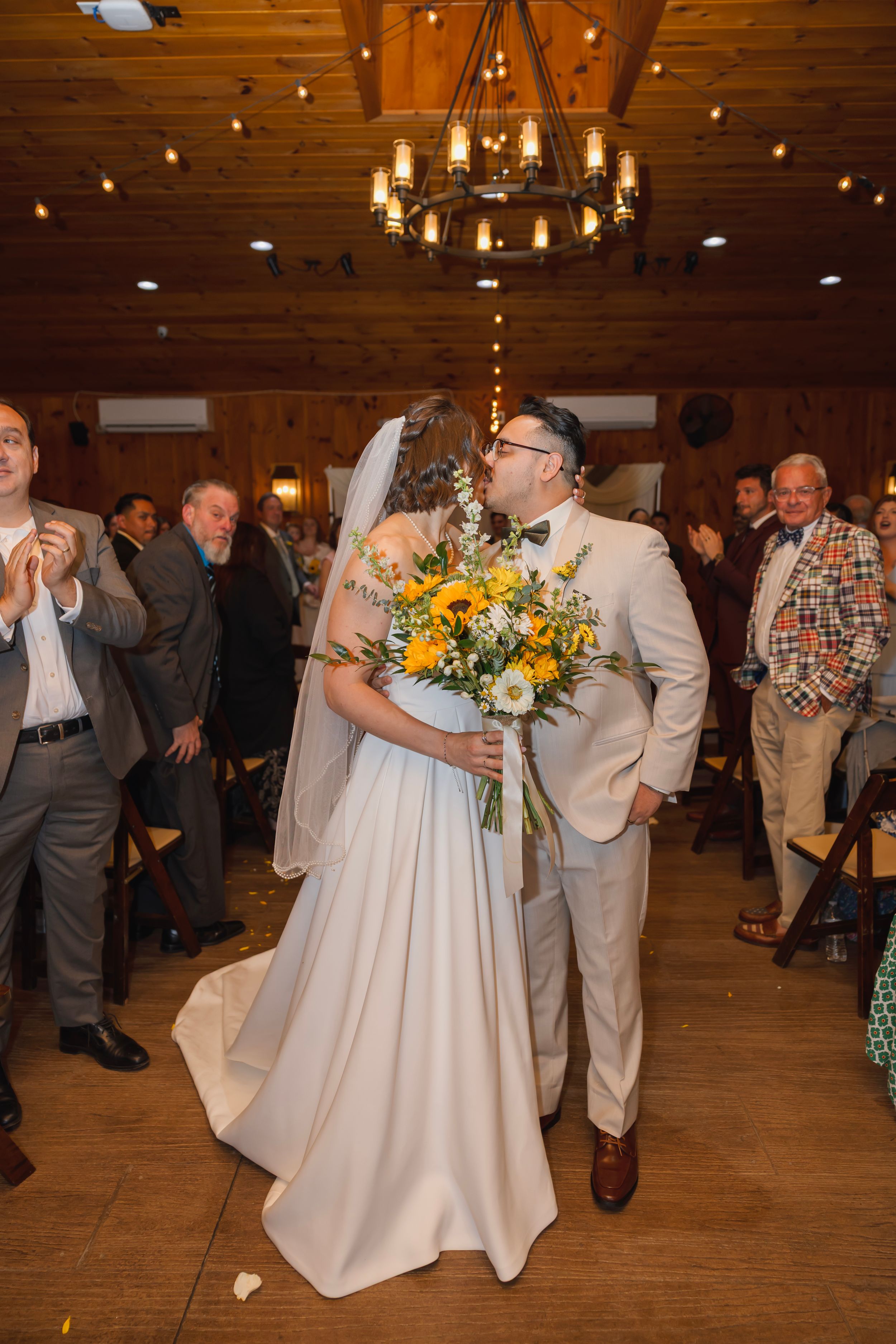 Newlyweds share a first kiss as husband and wife while walking down the aisle holding a sunflower bouquet.