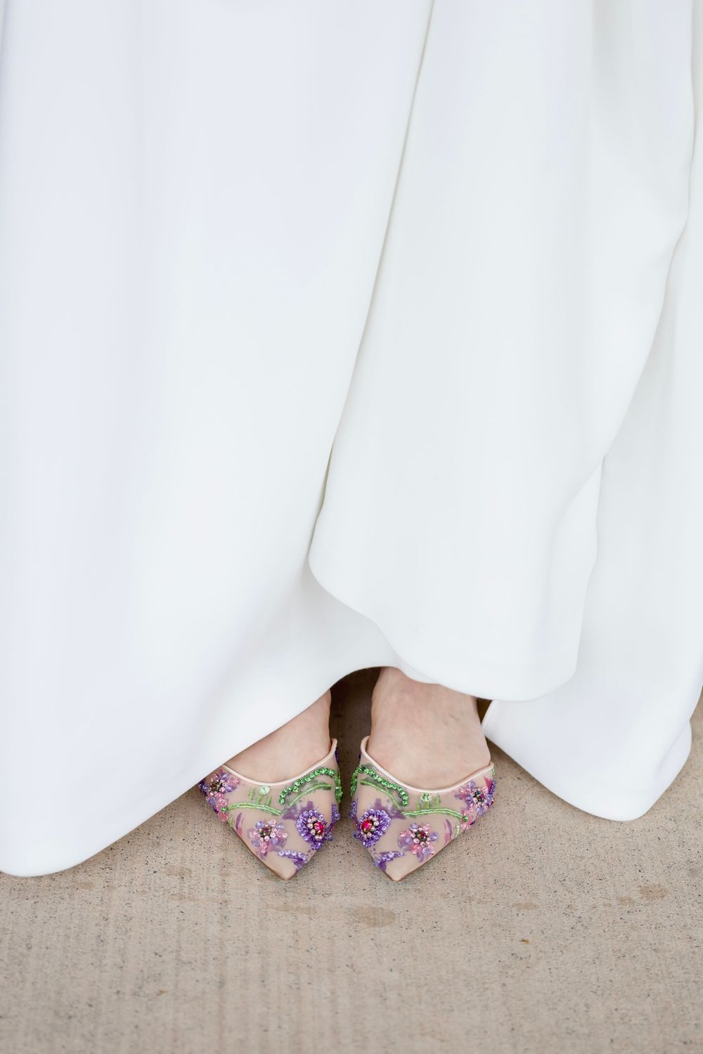 Bride wearing floral embroidered pointed-toe flats with purple flowers peeking beneath a white wedding gown.