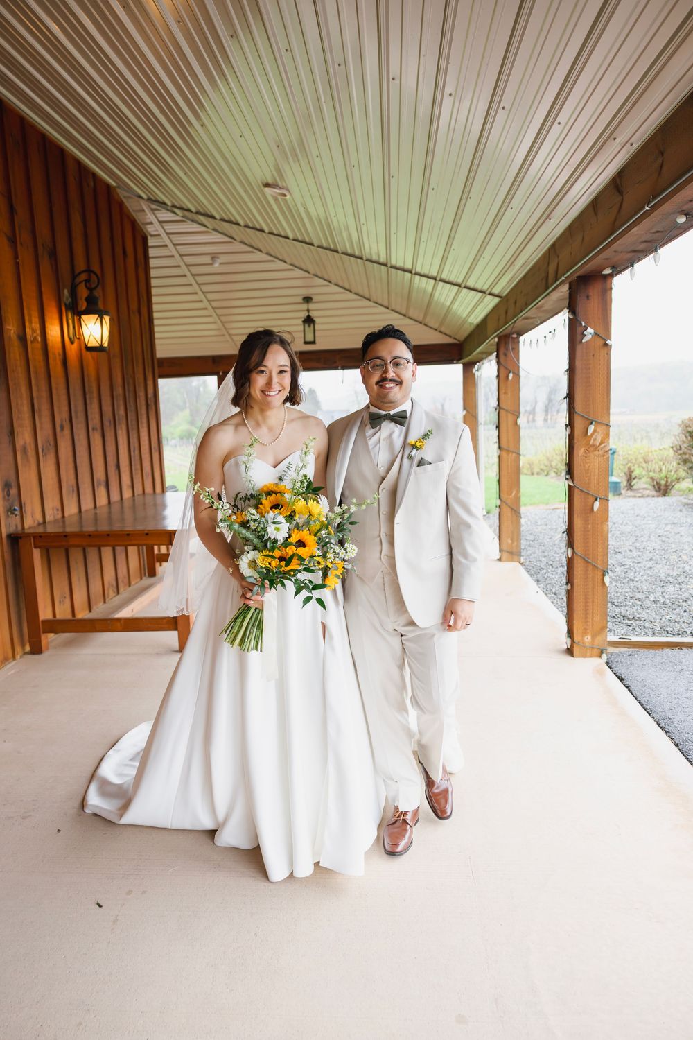 Bride holding sunflower bouquet and groom in cream suit walk together under wooden venue porch on their wedding day.