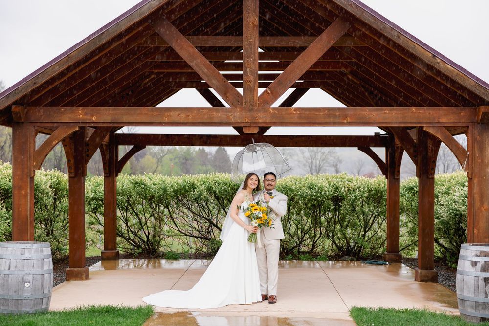 Bride and groom pose under a rustic wooden pavilion at a vineyard wedding venue, surrounded by lush greenery.
