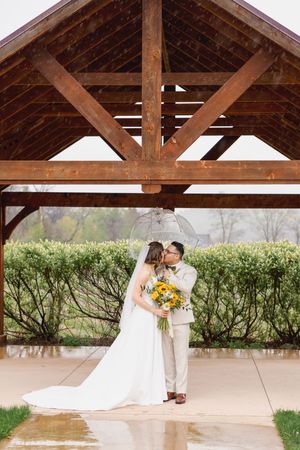 Bride and groom kissing under a rustic wooden pavilion on a rainy wedding day, holding a sunflower bouquet.