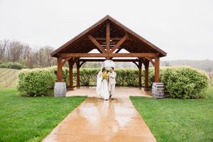 Bride and groom share umbrella under wooden pavilion on rainy wedding day at vineyard venue.