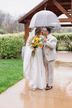 Bride and groom kissing under clear umbrella in rain, bride holding sunflower bouquet in white gown.