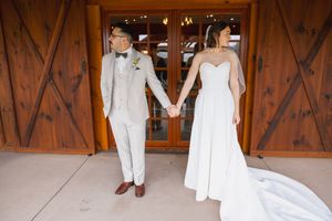 Bride and groom hold hands facing away from each other in front of rustic wooden barn doors on their wedding day.