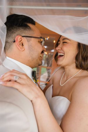 Bride and groom laughing together under wedding veil, bride wearing pearl necklace and engagement ring.