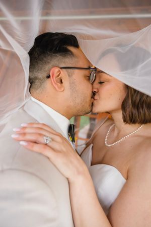 Newlyweds share a romantic kiss under the bride's veil, showcasing her diamond ring and pearl necklace.