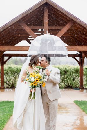 Bride and groom share a kiss under a clear umbrella in the rain, holding a sunflower bouquet outdoors.