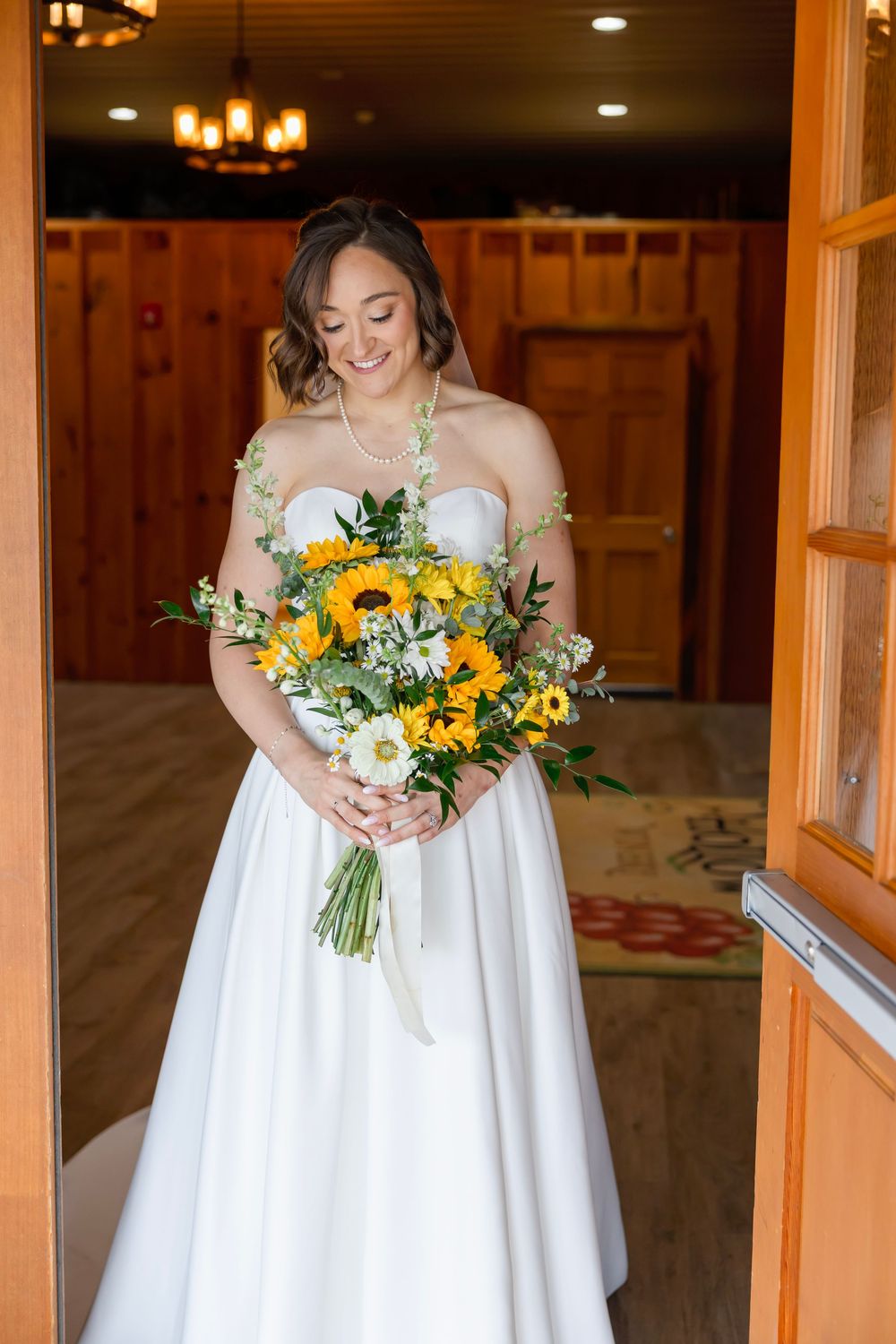Smiling bride in strapless white gown holding vibrant sunflower bouquet standing in wooden doorway.