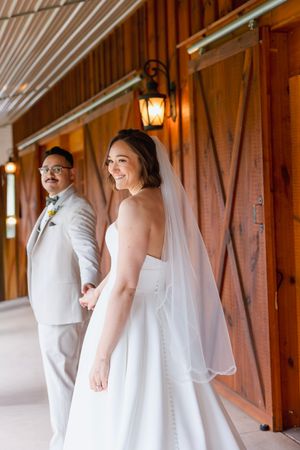 Bride in white strapless gown and veil holds grooms hand outside rustic barn wedding venue.