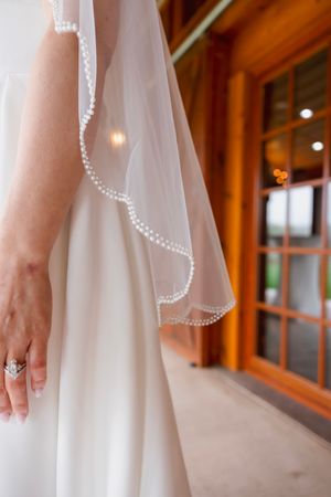 Close-up of pearl-trimmed bridal veil and diamond ring against white wedding gown near wooden door.