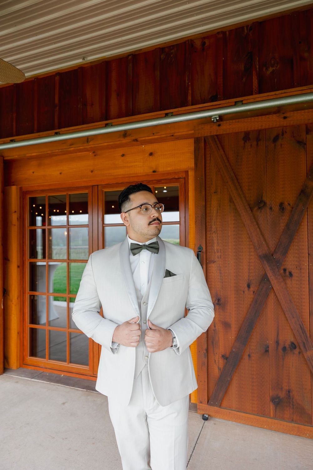 Groom in light gray tuxedo with bow tie stands confidently outside rustic barn wedding venue.