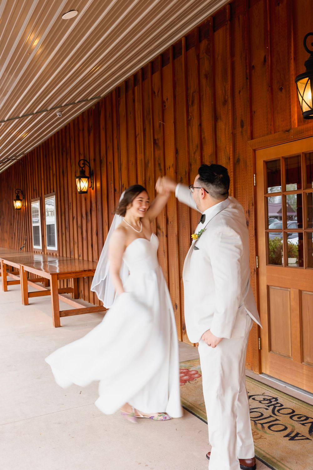 Bride and groom dancing on rustic barn venue porch, groom spinning bride in flowing white wedding dress.
