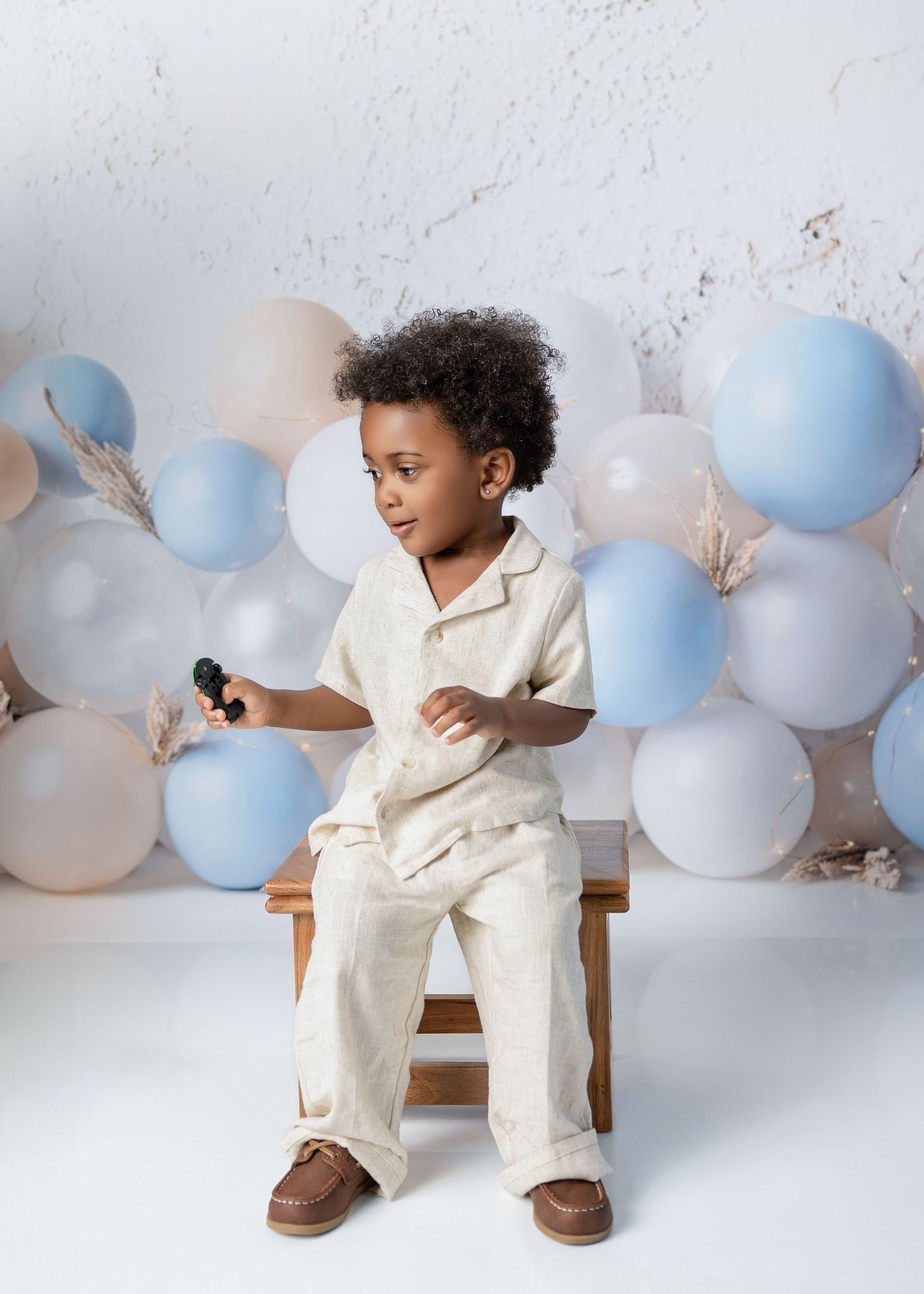 Young Black boy in cream linen outfit sits on wooden bench surrounded by blue and beige balloons.