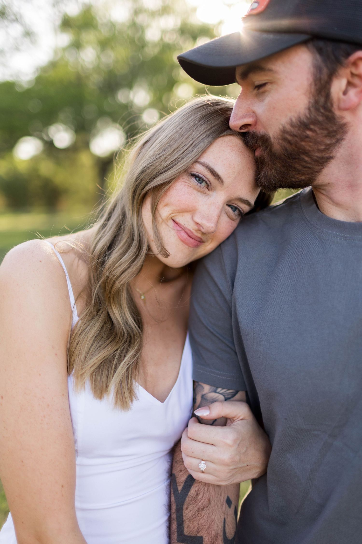 A couple shares a tender moment outdoors, man kissing woman's cheek as she smiles, wearing white dress and engagement ring in Austin, Texas.