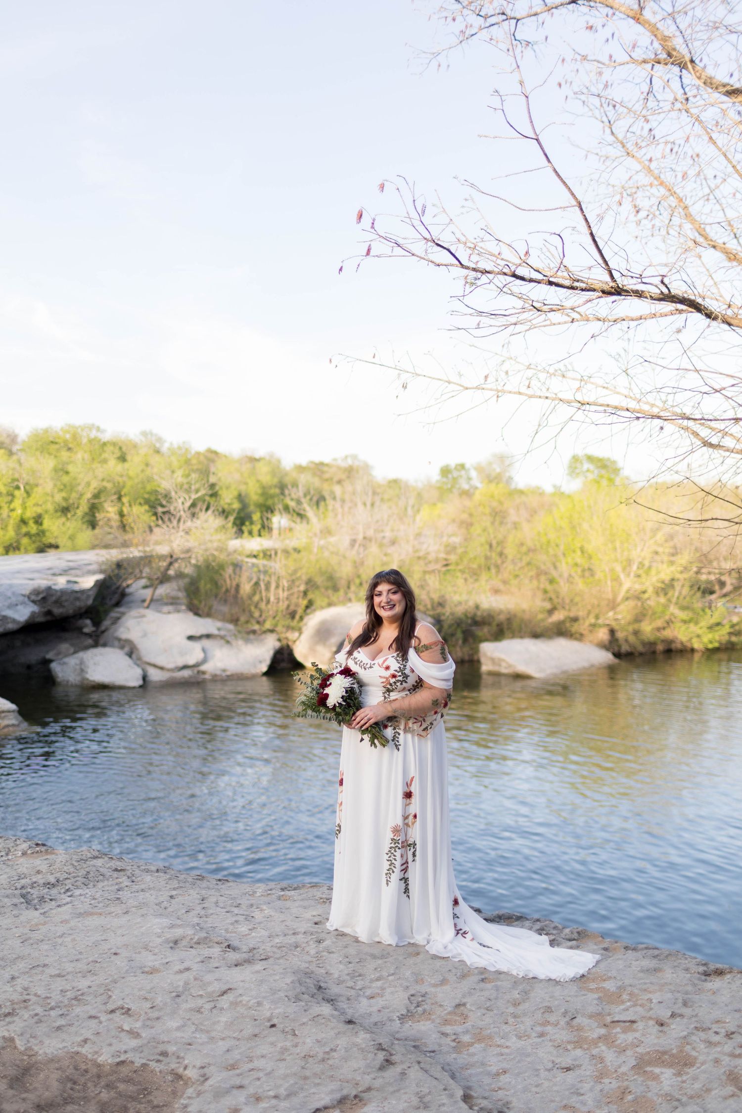 Bride in white floral embroidered gown twirling near a serene river, holding a bouquet outdoors.