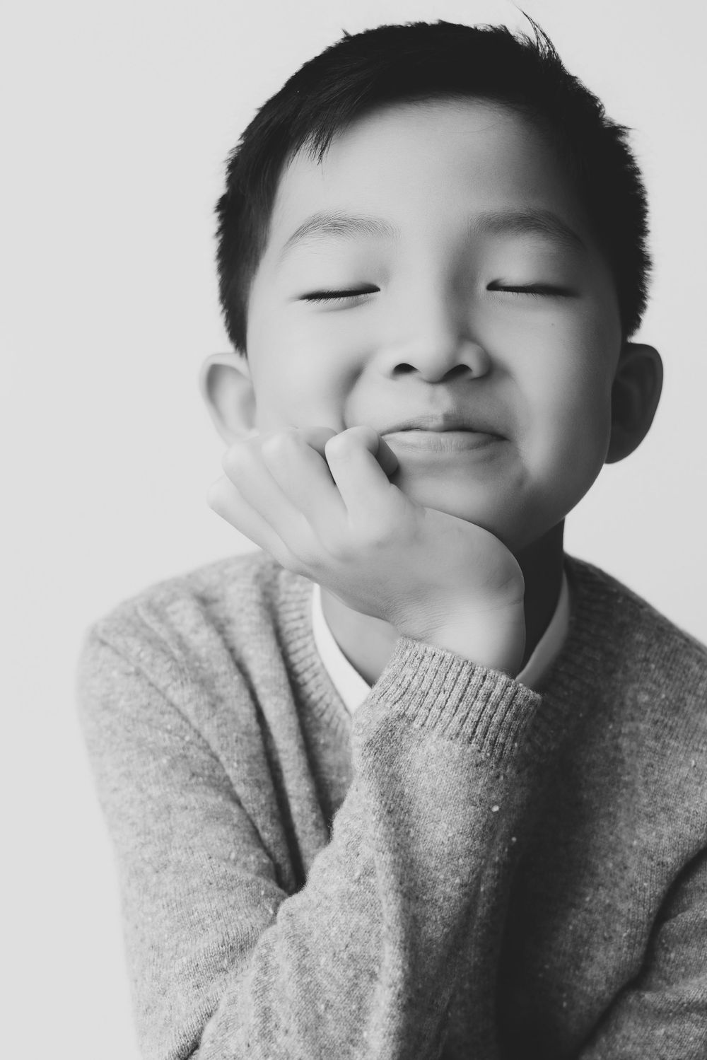 Black and white portrait of a smiling Asian boy with eyes closed, resting chin on hand, wearing a sweater.