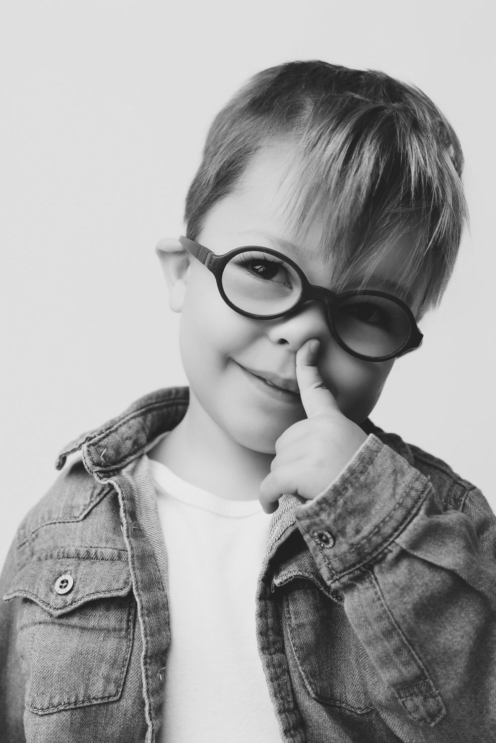Black and white photo of a cute child wearing oversized round glasses, smiling and pointing to nose, wearing denim jacket.