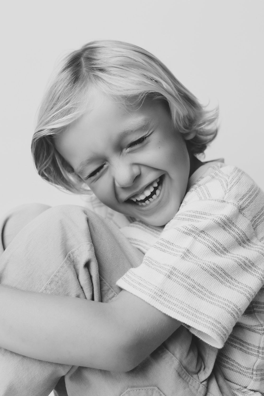 Black and white portrait of a laughing blonde child hugging their knees, radiating pure joy.