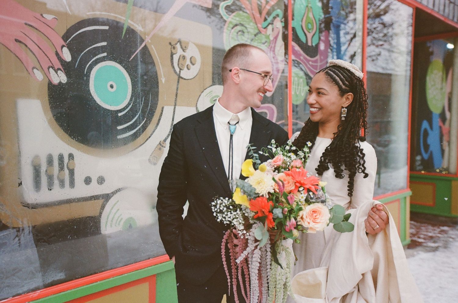 Couple in wedding attire smiling before colorful street mural, bride holding vibrant bouquet.