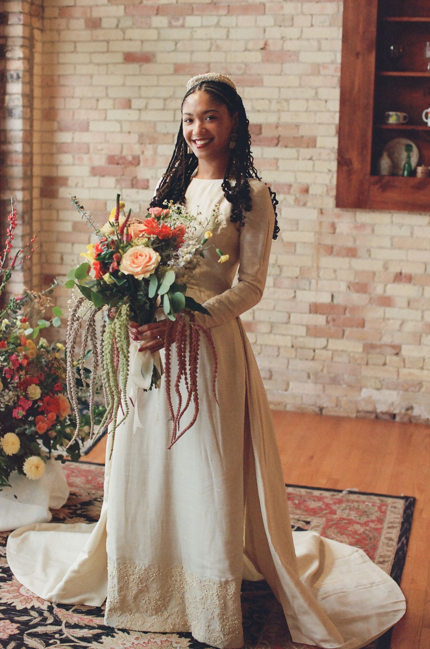 Smiling bride in ivory long-sleeve gown holding colorful bouquet with trailing greenery, standing in rustic brick-walled venue.