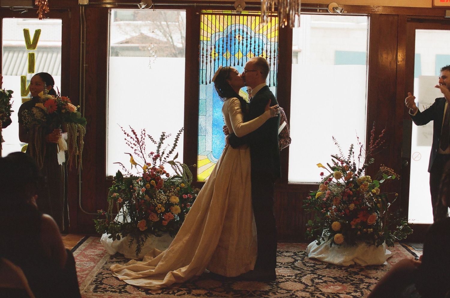 A bride and groom share their first kiss at an elegant indoor wedding ceremony surrounded by lush floral arrangements.