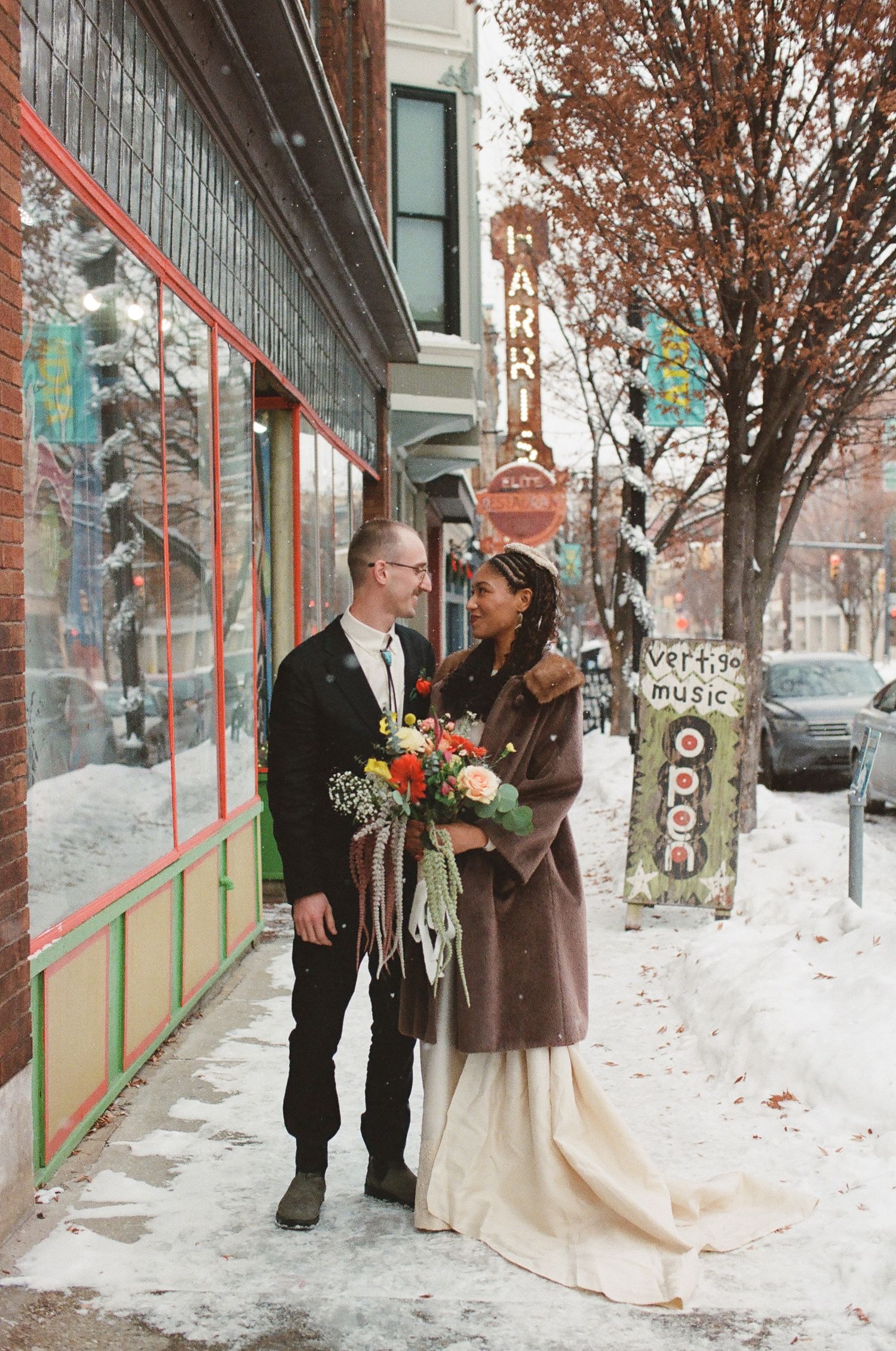 Couple in wedding attire stands on snowy urban sidewalk outside Harris music store, bride holding colorful bouquet in winter scene.