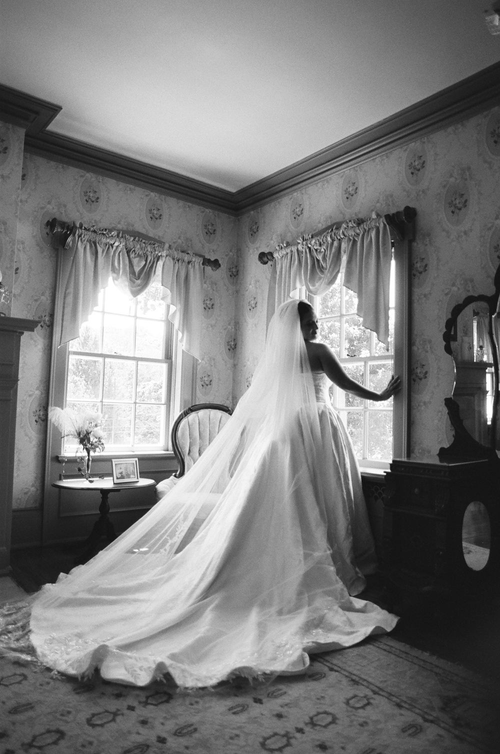 Black and white photo of a bride in a flowing gown and veil standing by a window in a vintage Victorian room.