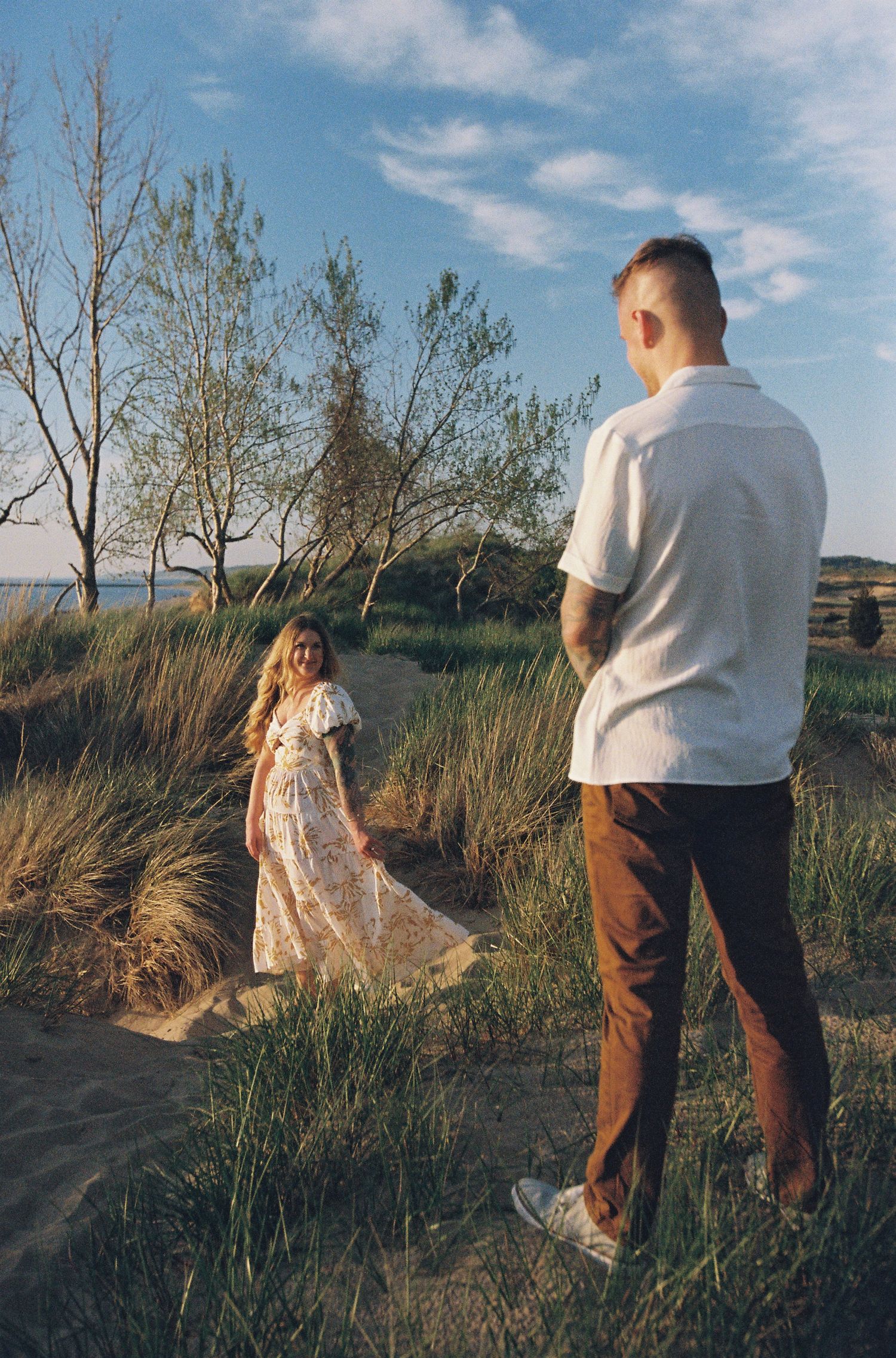 Couple standing on sandy dunes near trees and water at golden hour, woman in floral dress smiling at man in white shirt.
