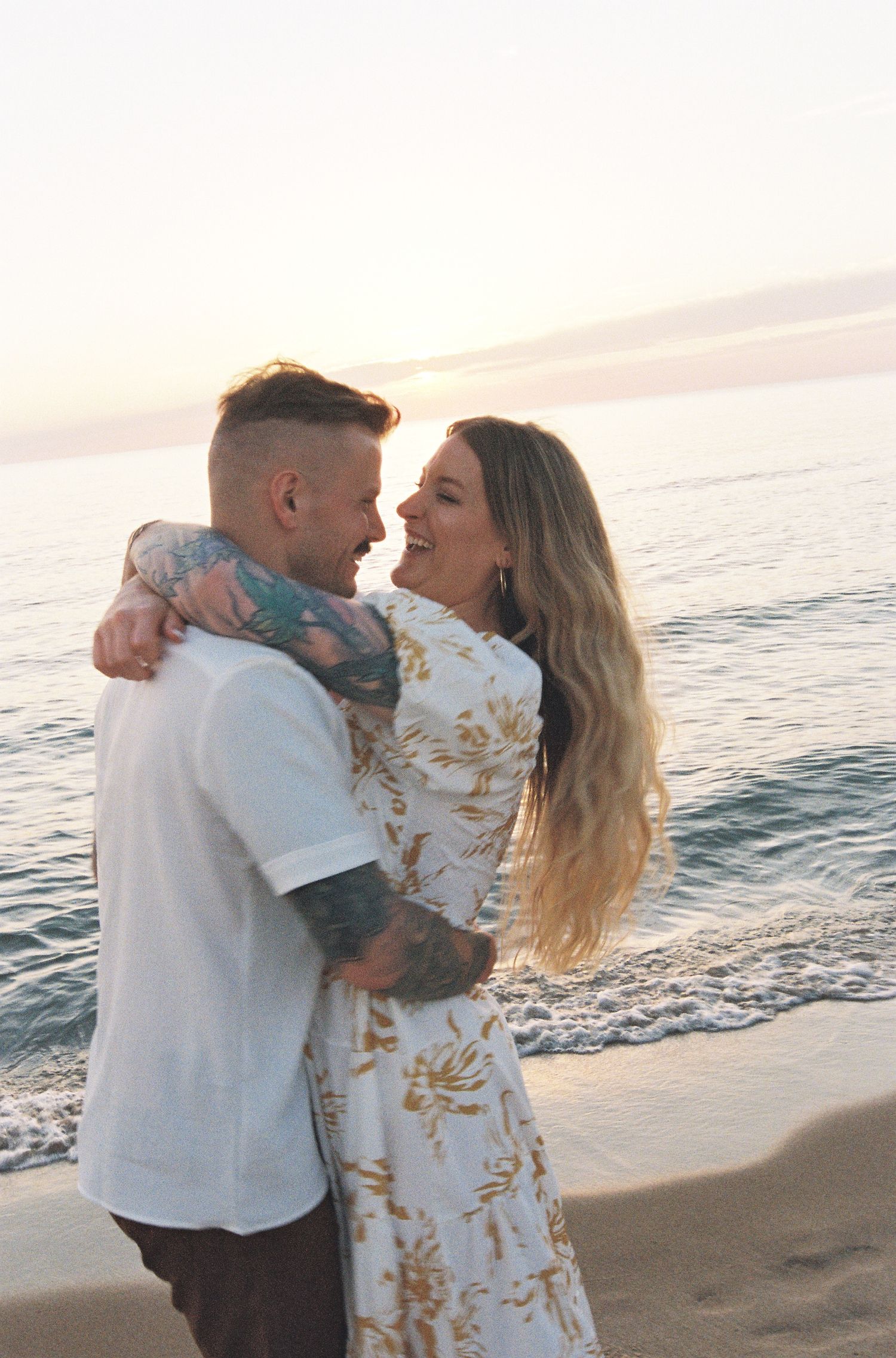A couple embraces and smiles at each other on a beach at sunset, man with tattoos in white shirt, woman in floral dress.