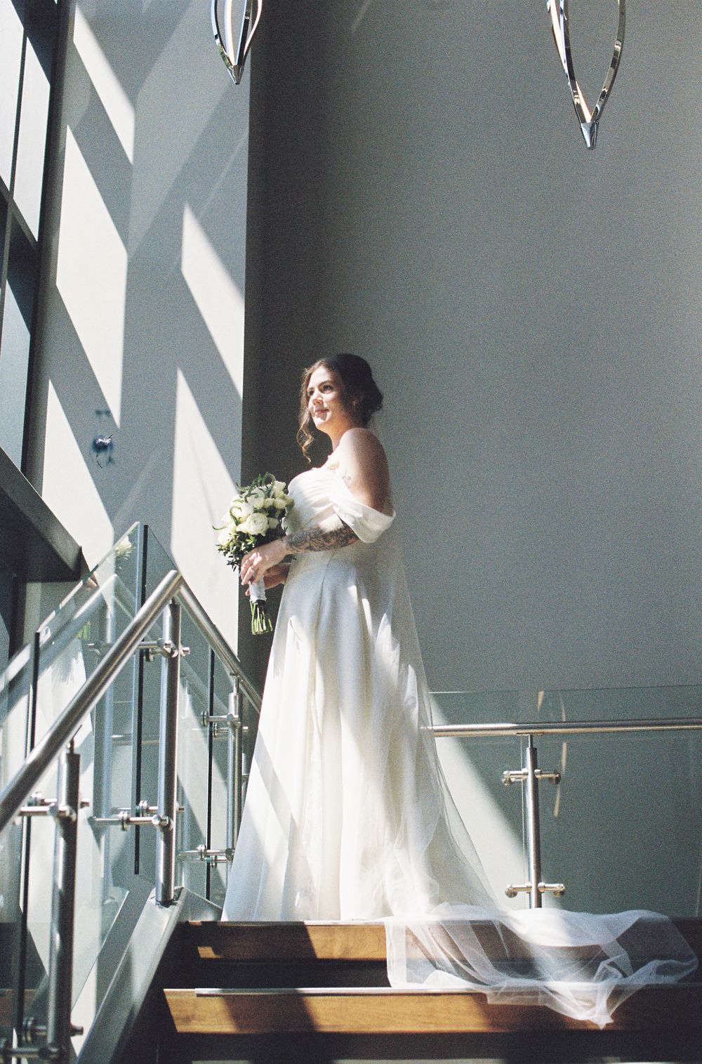 Bride in elegant white off-shoulder gown holding bouquet stands on sunlit staircase with glass railings.