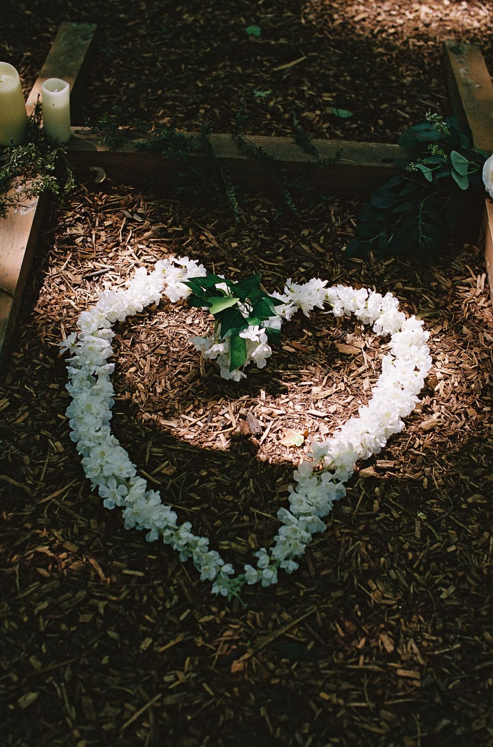 White flower petals arranged in a heart shape on mulch, illuminated by soft candlelight in a romantic outdoor wedding setting.