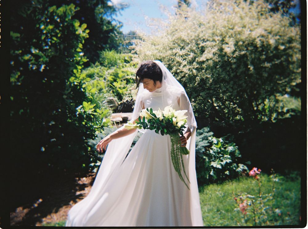 Bride in white gown and veil holding floral bouquet in lush green garden on wedding day.
